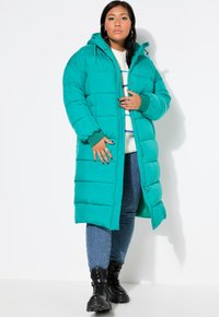 Teal pufferjakke med hætte, der har quiltede sektioner, frontlynlås og elastiske manchetter. Båret over en hvid sweater og blå jeans med dekorationer.