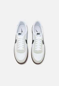 Tenisky Nike s bílým koženým svrškem, šedými semišovými akcenty, černým logem swoosh a hnědou gumovou podrážkou; tkaničky jsou bílé.