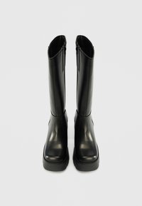 Melnas ceļgala augstuma zābaki, kas izgatavoti no gludas ādas ar noapaļotu pirkstu un biezu gumijas zoli. Iekšpusē ir rāvējslēdzējs, kas nodrošina ērtu uzvilkšanu.