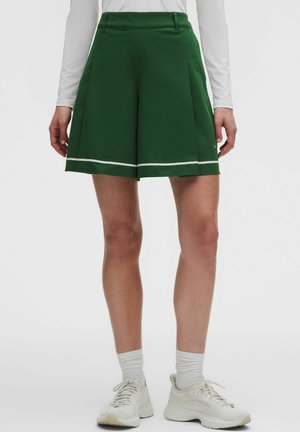 Shorts verts plissés avec bordure blanche portés avec des chaussettes blanches et des chaussures de sport blanches, montrés de la taille jusqu’aux pieds sur une personne.
