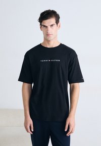 Camiseta de algodón negra con mangas cortas, que presenta el logo de "TOMMY HILFIGER" en blanco y rojo, cuello redondo y fit relajado.