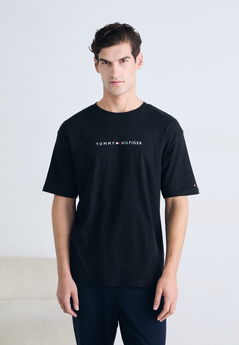 Camiseta de algodón negra con mangas cortas, que presenta el logo de "TOMMY HILFIGER" en blanco y rojo, cuello redondo y fit relajado.