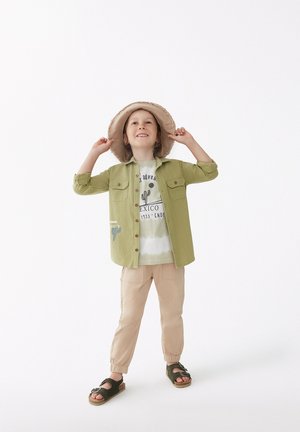 Niño vestido con una camisa verde de botones, una camiseta blanca con gráfico, pantalones jogger beige y sandalias oscuras, acompañado de un sombrero de paja claro.