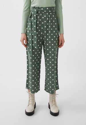 Green cropped pants with white polka dots and belt, paired with light green long-sleeve top and white lace-up ankle boots.