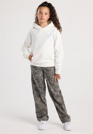 Meisje met krullend haar in een witte hoodie en een broek met luipaardprint staat met één hand in de zak tegen een effen achtergrond.