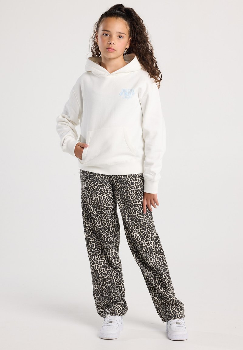 Girl with curly hair in a white hoodie and leopard-print pants stands with one hand in pocket against a plain background.