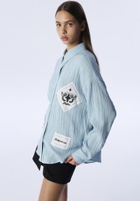 Jasnoniebieska, teksturowana koszula z długim rękawem w oversize'owym kroju. Posiada białe łatki z czarnymi wzorami motyli i tekstu. Krótkie rękawy, styl casual.