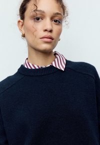 Temno modra jopica z teksturiranim pletenjem, z rebrastim izrezom, nošena čez srajco z ovratnikom v rdeče-belem črtastem vzorcu.