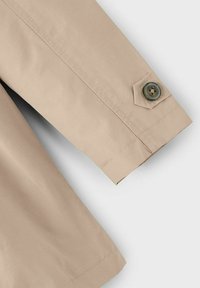 Manche en tissu beige avec des coutures visibles et un revers ornée d'un bouton verdâtre cousu sur une petite languette.