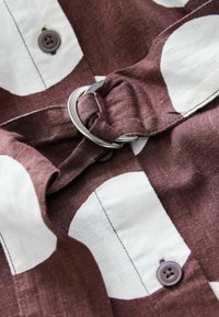 Pruun ja valge mustriga särgikangad, millel on metallringi detail ja tumedad nööbid, demonstreerides lina sarnast tekstuuri ja vaba stiili.