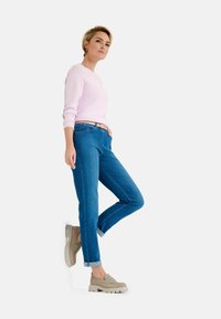 Pull rose clair, jeans en denim bleu, et chaussures beige à semelles épaisses ; coupe décontractée avec des manches retroussées et un détail de ceinture.