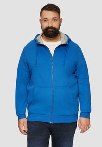 Niebieska rozpinana bluza z miękkiego materiału, z szarym podszyciem kaptura, dwoma przednimi kieszeniami i regulowanymi sznurkami. Nosi się ją na białej koszuli.