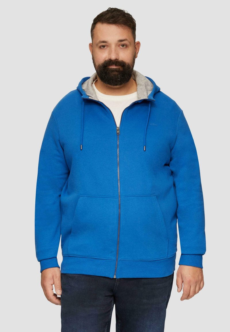 Niebieska rozpinana bluza z miękkiego materiału, z szarym podszyciem kaptura, dwoma przednimi kieszeniami i regulowanymi sznurkami. Nosi się ją na białej koszuli.