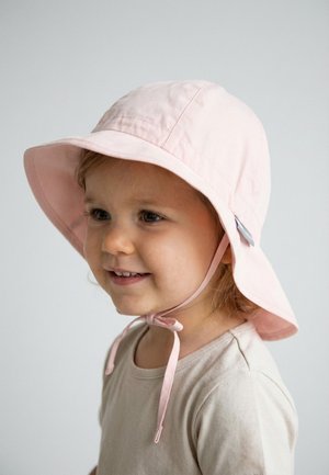 Young child smiling, wearing a light pink sun hat with chin straps tied, and a beige shirt against a plain light background.