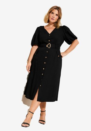 Kvinde med skulderlangt hår iført en sort midi-kjole med pufærmer, guldknapper, bælte i taljen og sorte hæle med ankelrem.