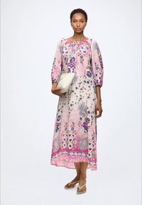Mujer con vestido midi de manga larga con flores en rosa y púrpura, sosteniendo un bolso clutch blanco y llevando sandalias blancas sobre un fondo blanco.