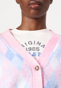 Cárdigan en patrón de rombos rosa y azul, con escote en v, cierre de botones y superpuesto sobre una camiseta gráfica blanca con textura acanalada.