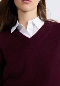 Bordowy sweter w serek, wykonany z miękkiego dzianiny, noszony na białej koszuli z kołnierzykiem. Posiada prążkowane detale przy mankietach i dekolcie.