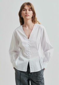 Camicia bianca con colletto a V, maniche lunghe a palloncino e finitura testurizzata. Presenta una fila di bottoni lungo la parte anteriore e un orlo curvo.