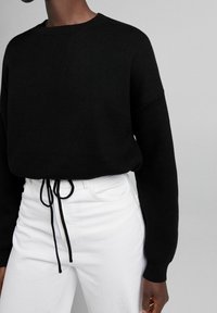 Schwarzer Strickpullover mit rundem Halsauschnitt und überschnittenen Schultern, kombiniert mit weißen, hochgeschnittenen Hosen mit einem Kordelzug an der Taille.