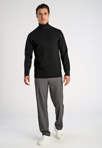 Schwarzer Rollkragenpullover aus glattem Stoff, kombiniert mit grauen Hosen und weißen Sneakers, die einen modernen, lässigen Look kreieren.