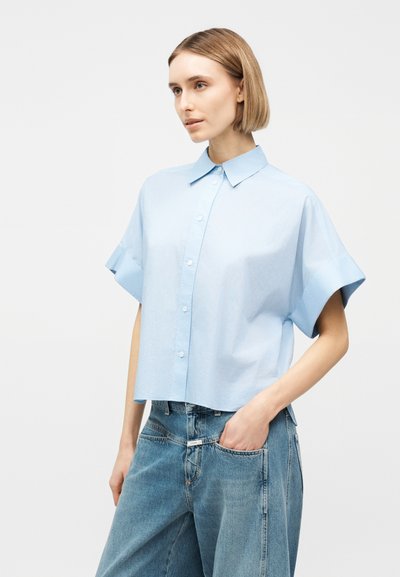 Γυναίκα με κοντά ξανθά μαλλιά, φορώντας ένα ανοιχτό μπλε πουκάμισο με κοντά μανίκια και χαλαρά μπλε τζιν, με το χέρι στην τσέπη, κοιτάζοντας μπροστά.