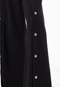 Pantalones negros con una textura suave, que cuentan con un cierre lateral de botón con botones dorados brillantes y un corte relajado.