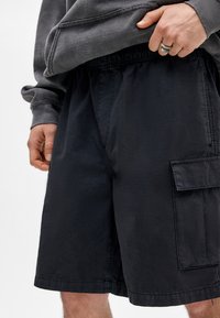 Shorts cargos noirs en tissu léger, avec une taille élastique, une poche cargo sur le côté et une coupe décontractée. Fini texturé.