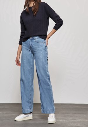 Osoba ubrana w granatowy sweter z długim rękawem o fakturowanej strukturze, jasnoniebieskie dżinsy z wysokim stanem i szerokimi nogawkami oraz białe trampki, stojąca z jedną ręką na biodrze.