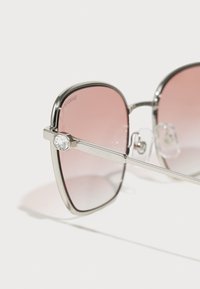 Ochelari de soare Swarovski cu ramă argintie, lentile roz nuanțate și un accent decorativ din cristal pe brățară. Design unghiular și geometric.