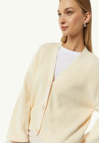 Helle gelbe Strickjacke mit V-Ausschnitt, langen Ärmeln und vier Knöpfen. Die Textur ist gerippt und weich.