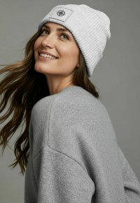 Light gray ribbed knit beanie with a compass logo patch; soft texture, relaxed fit. Worn with a gray sweater showcasing a cozy design.
