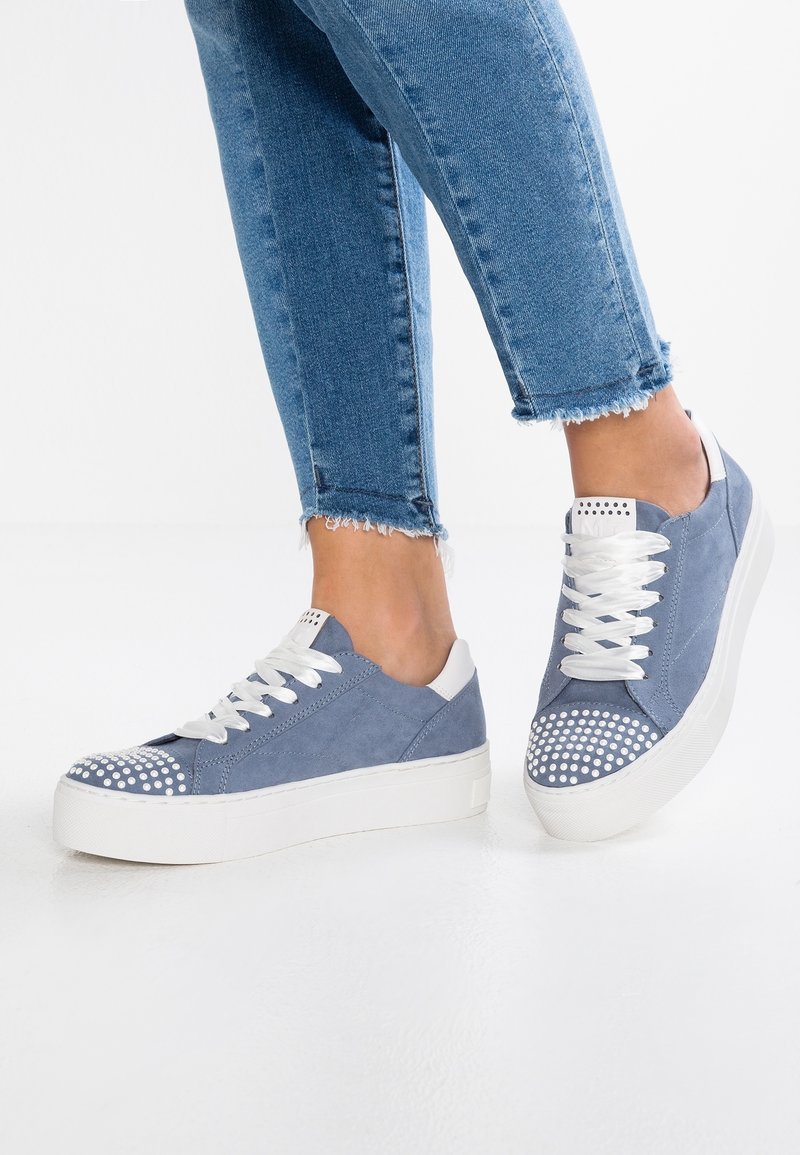 Personne portant des baskets en daim bleu avec des bouts blancs cloutés et des semelles blanches épaisses, assorties à un jean bleu à ourlet effiloché.