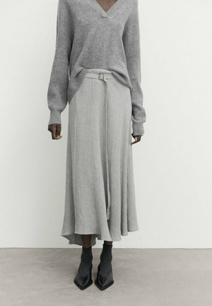 Personne portant un pull gris à col en V, rentré dans une jupe midi fluide ceinturée, avec des bottines noires et des chaussettes, se tenant sur un sol gris.