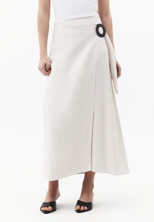 Biała midi spódnica kopertowa z czarną okrągłą klamrą, noszona z białym topem bez rękawów i czarnymi sandałami na obcasie z odkrytymi palcami.