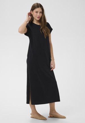 Jeune femme aux longs cheveux portant une robe midi noire à manches courtes avec une fente latérale et des chaussures plates tissées beiges, debout et se touchant les cheveux.