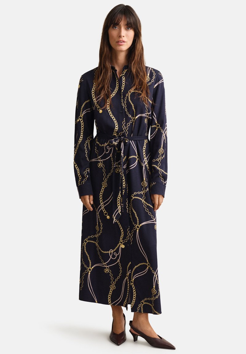 Femme portant une longue robe bleu marine avec des motifs de chaînes dorées, devant boutonné, ceinture à la taille, manches longues, et chaussures basses à talons pointus foncées.