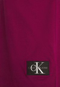 Parche rectangular negro con el logo de Calvin Klein Jeans sobre un fondo de tela magenta oscuro.