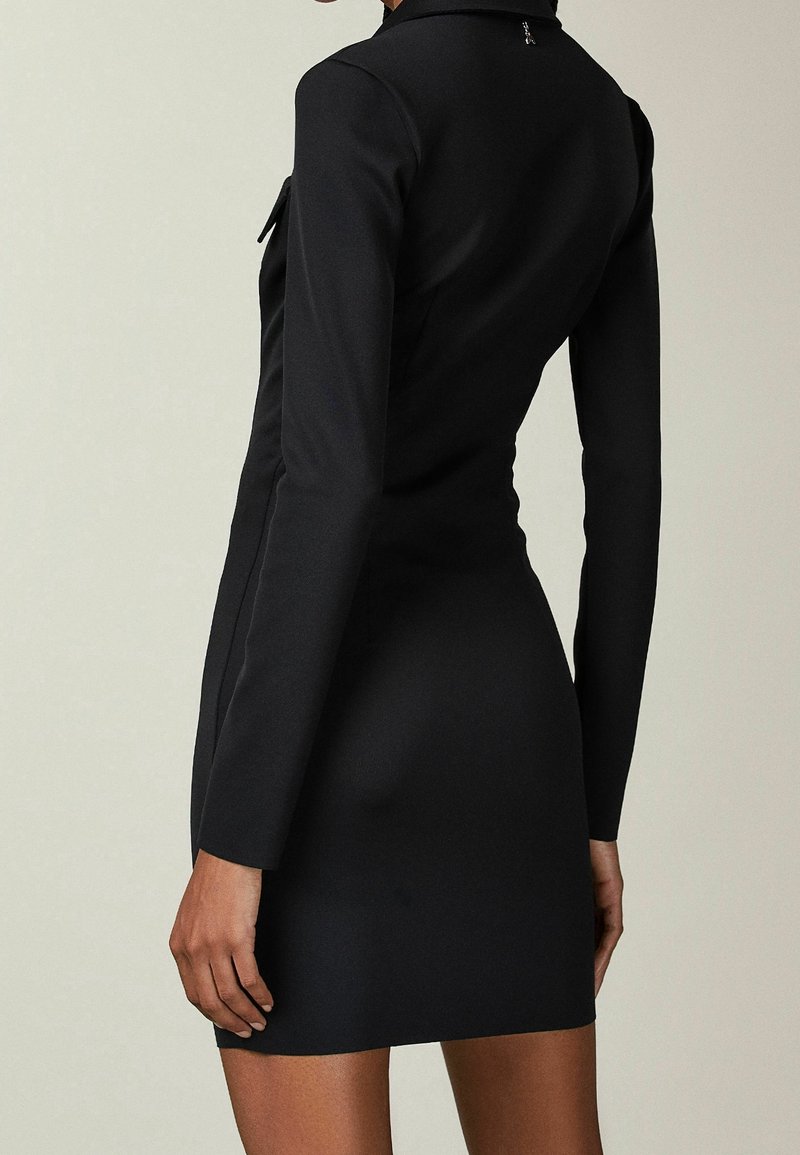 Femme portant une robe noire cintrée à manches longues, debout dos à la caméra sur un fond uni.