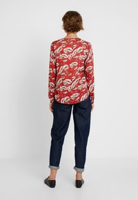 Rode blouse met lange mouwen en bloemenpatronen, gecombineerd met opgerolde donkere spijkerbroek. Schoenen zijn zwarte ballet flats, bekeken vanaf de achterkant.