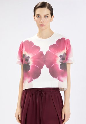 Donna che indossa una maglietta bianca a maniche corte con grandi stampe floreali simmetriche rosa e viola, abbinata a pantaloni larghi color marrone scuro.