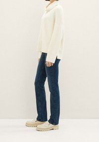 Personne portant un pull à col roulé crème, un jean droit bleu foncé et des mocassins beiges épais, debout de profil sur un fond uni.