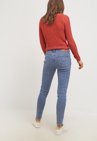 Roter strukturierten Pullover kombiniert mit blauen Skinny-Jeans, die ausgeprägte Gesäßtaschen und einen Logo-Patch aufweisen; abgerundet mit weißen Sneakers.
