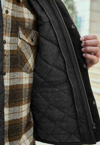Quilted black jacket with brown and beige plaid lining. Features a chest pocket and zip closure; smooth texture and contrasting patterns.