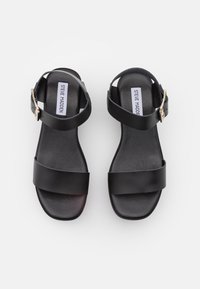 Sandalias de cuero negro con dos correas anchas y una correa en el tobillo, que presentan una hebilla de tono dorado. Suela plana con una textura suave.
