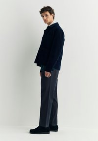 Marineblauwe ribfluwelen jas met twee voorzakken, gedragen over een groen overhemd, gecombineerd met donkergrijze broek en zwarte suède schoenen.