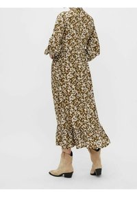 Kvinna klädd i en brun blommig midiklänning med långa ärmar och beigea mocka ankelskor, stående mot en enfärgad ljus bakgrund.