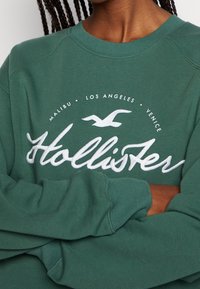 Vihreä collegepaita py öreällä pääntiellä, jossa on valkoinen kirjailtu logo, jossa on lintu ja teksti: "Hollister, Malibu, Los Angeles, Venice."