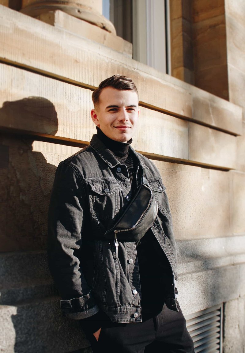 Black denim jacket over a black turtleneck, accessorized with a black crossbody bag. The background features a textured stone wall.