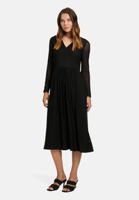 Zwarte midi-jurk met lange doorschijnende mouwen, V-hals en een geraffineerde taille. Soepele textuur; gedragen met zwarte slide sandalen.